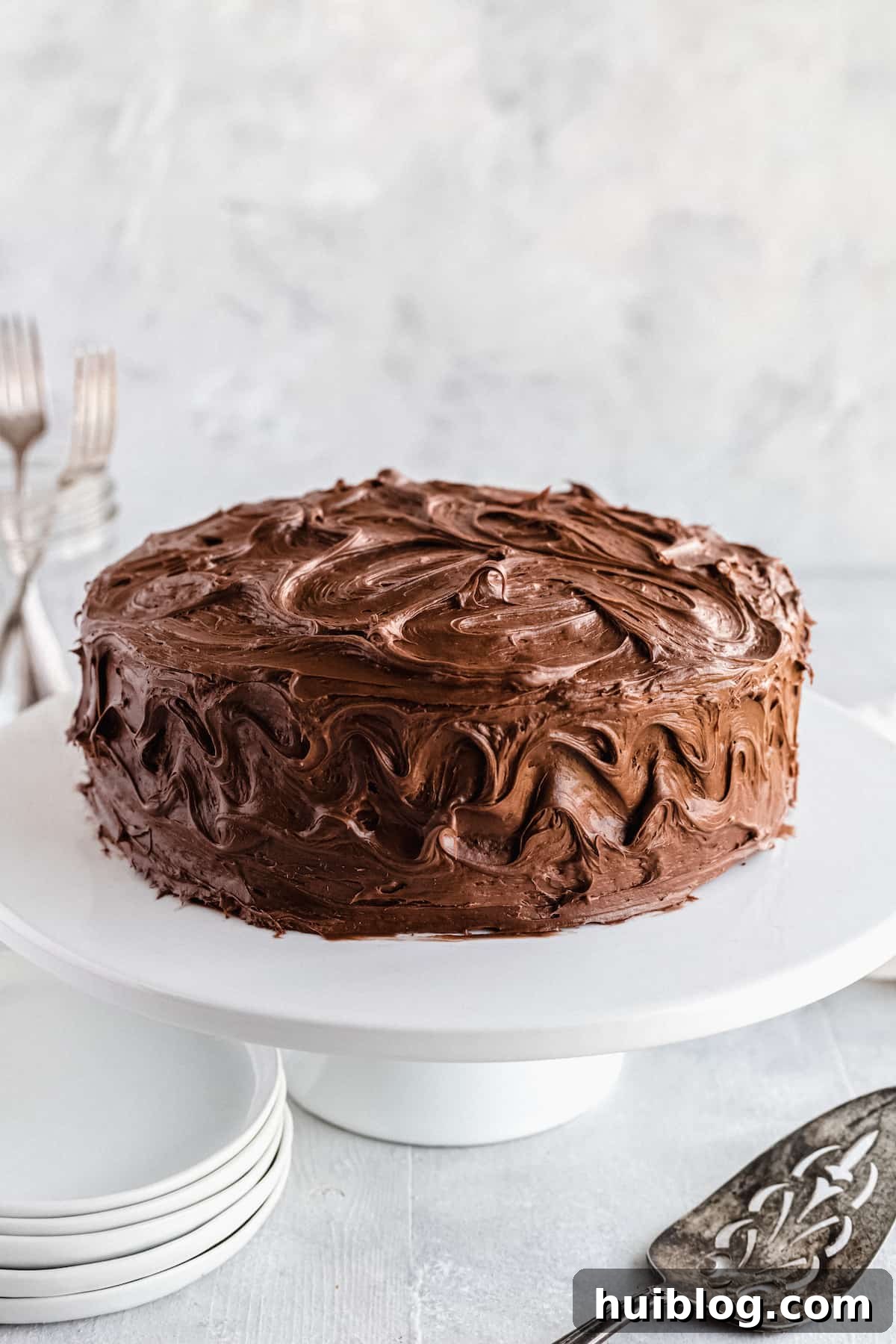 A beautifully frosted chocolate cake on a white cake stand, with a dessert spatula nearby