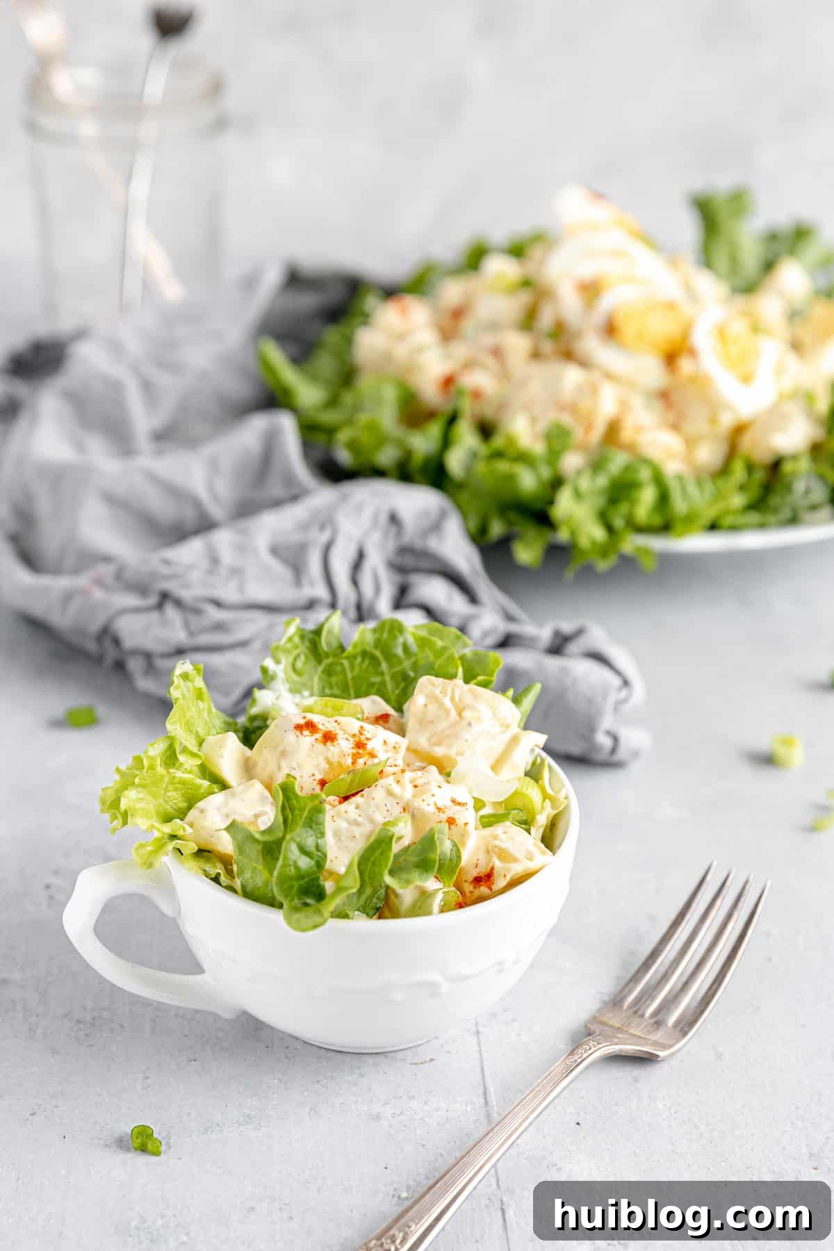 All-American Potato Salad 3 A Serving of Potato Salad in a Mug with the Remaining Salad on a Plate in the Background, showing off its creamy texture and visible ingredients.