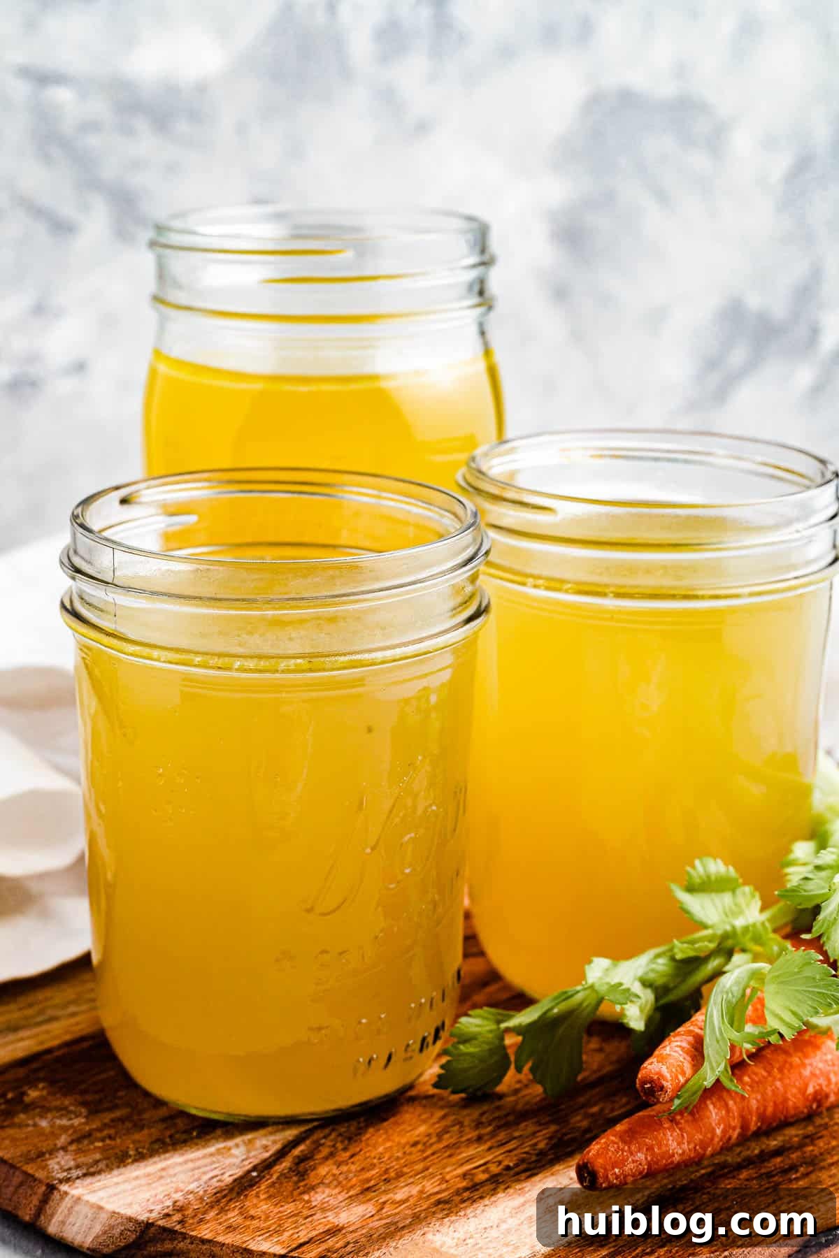 Liquid Gold: Instant Pot Chicken Broth 2 Three jars of homemade chicken broth on a cutting board with raw carrots nearby, symbolizing freshness and quality.
