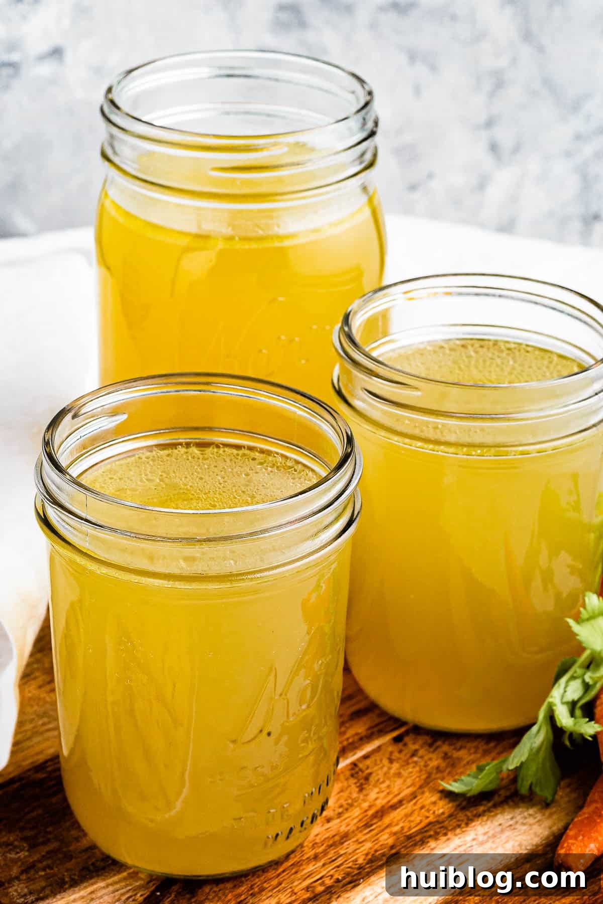 Liquid Gold: Instant Pot Chicken Broth 7 Three jars of chicken broth on a cutting board with raw carrots positioned next to them, emphasizing the fresh, natural ingredients.