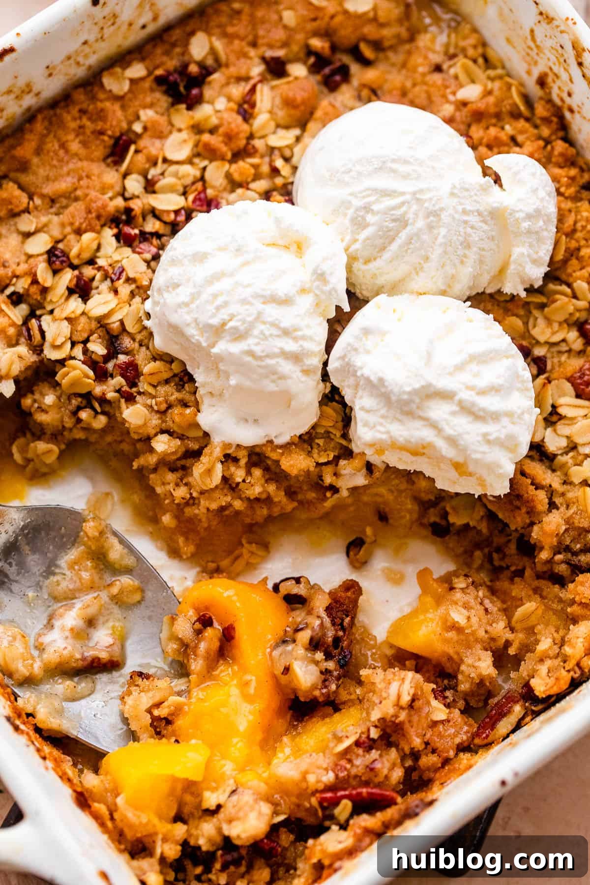 A close-up shot of a peach crisp baking dish, with melting vanilla ice cream scoops cascading over the golden-brown, bubbly topping and sweet peach filling.
