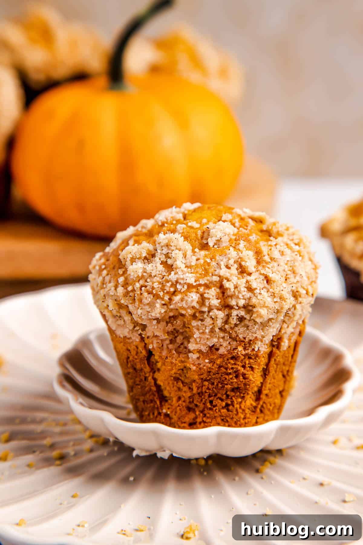 A single, perfectly baked pumpkin muffin on a pristine white plate, with a soft focus on three more muffins in the background, all adorned with a golden cinnamon crumble topping.