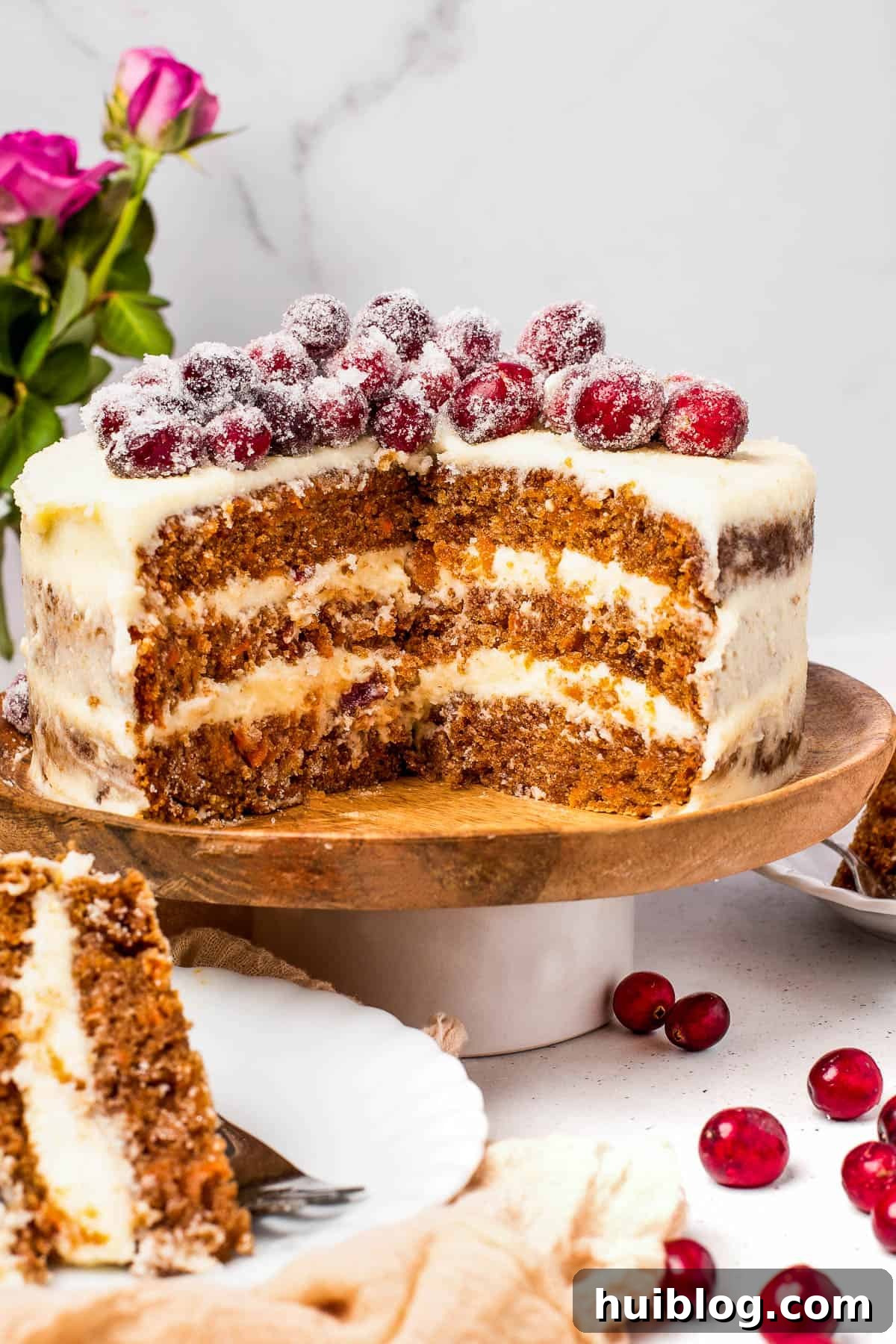 A three-layer carrot cake on a cake stand, with two slices cut out and set on dessert plates nearby, in a festive setting.