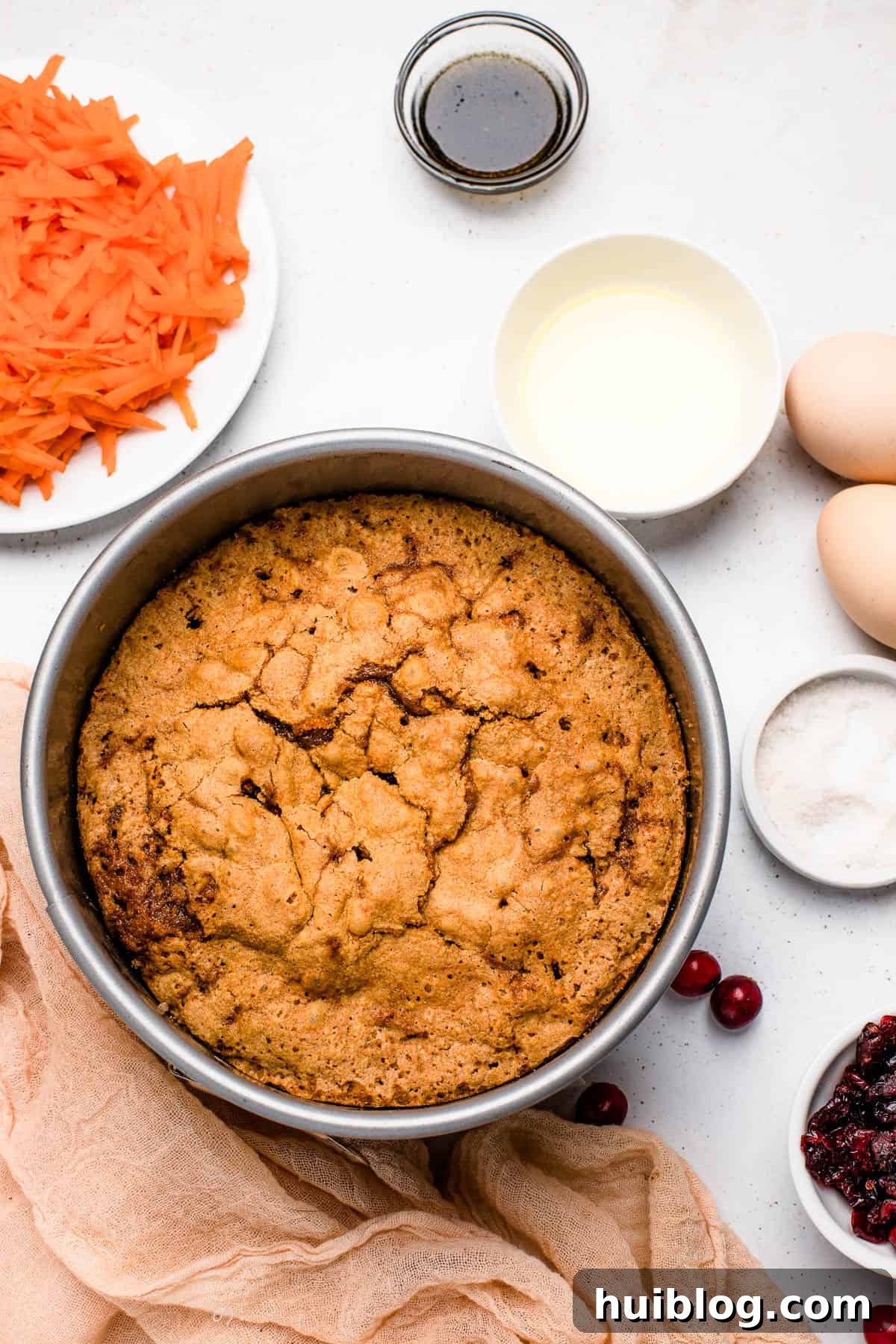 A baked cranberry carrot cake in the baking pan, cooling on a countertop with small dishes of ingredients nearby, showcasing the rustic appeal.