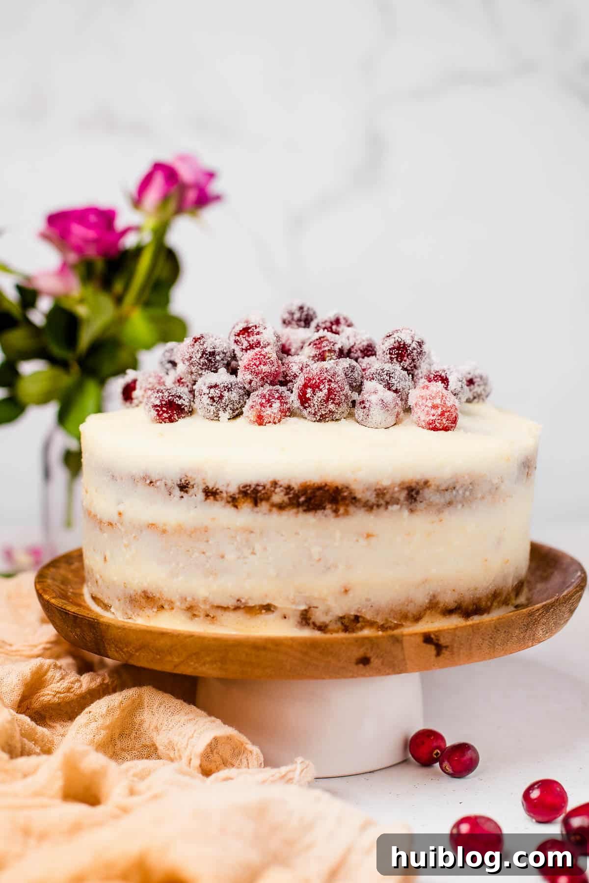 An assembled and iced cranberry carrot cake on a cake stand, generously topped with glistening sugared cranberries and a sprig of rosemary.