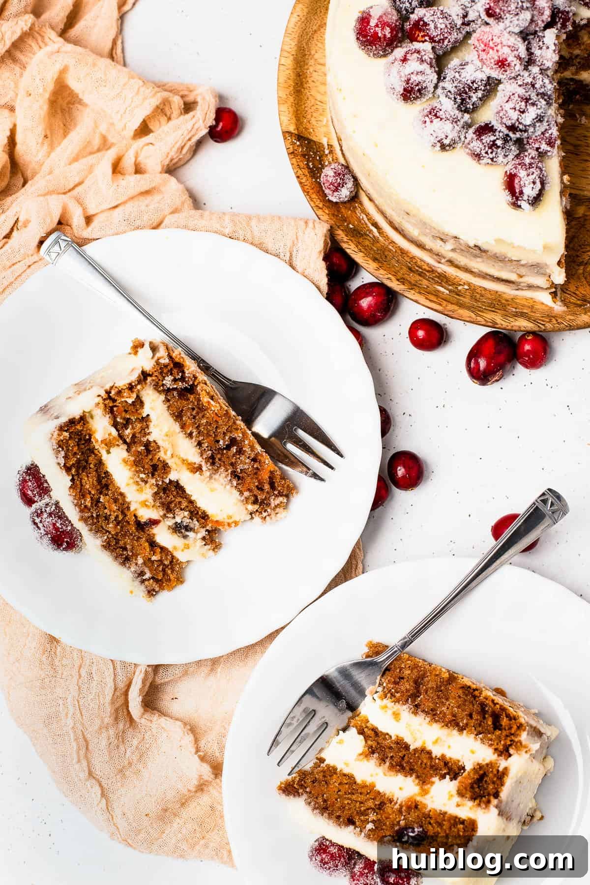 Slices of sugared cranberry layer cake on dessert plates, next to the remaining cake on a cake stand, decorated with fresh cranberries and rosemary.