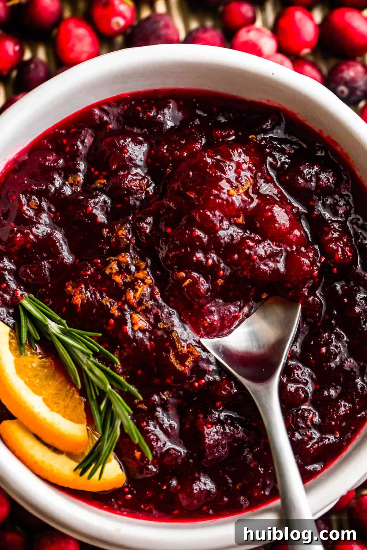 Overhead close-up shot of a spoon inside a bowl filled with homemade cranberry sauce, garnished with a rosemary sprig and fresh orange slices.