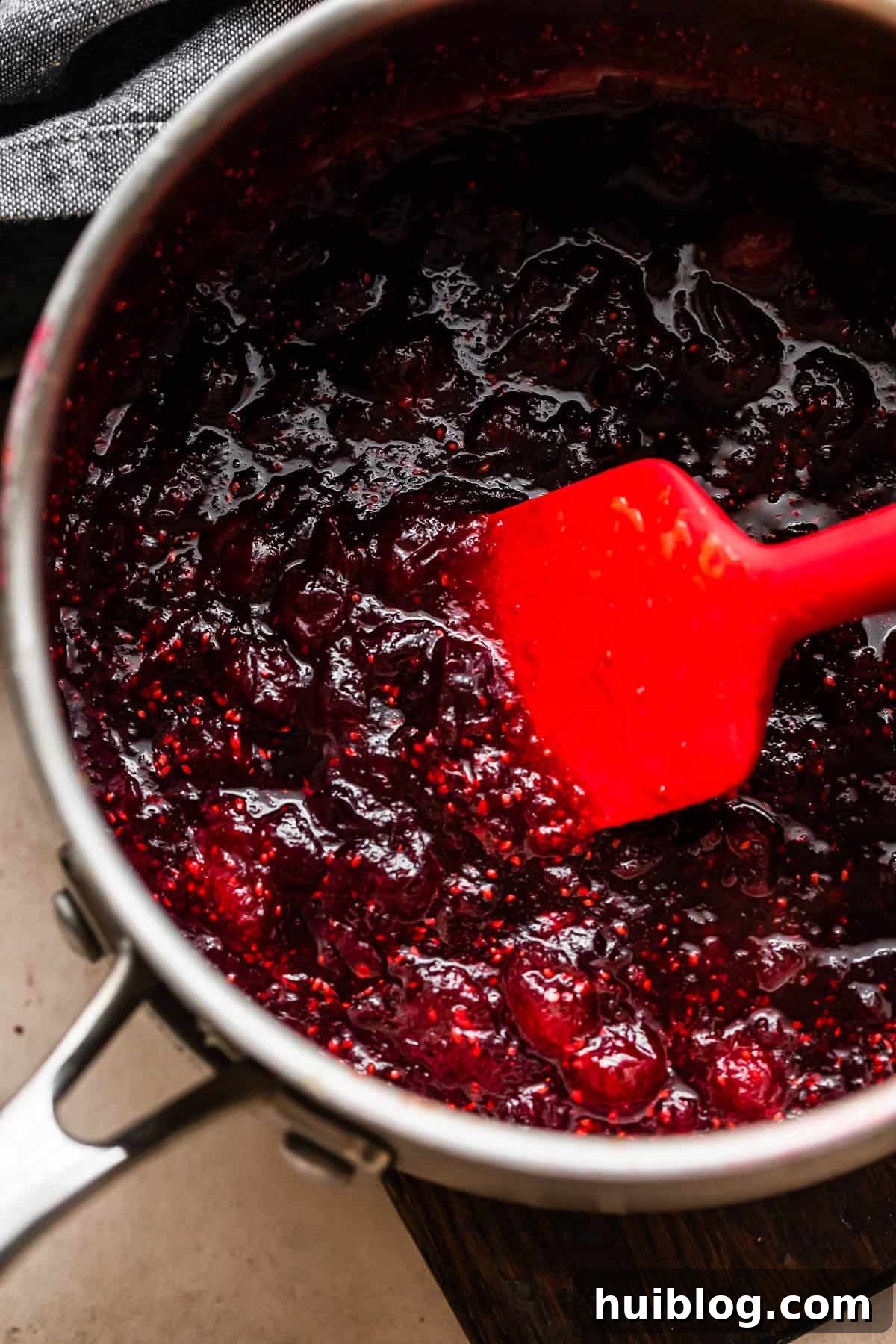 Cooking cranberry sauce in a saucepan, being stirred with a red rubber spatula, showing the cranberries bursting and the sauce thickening.