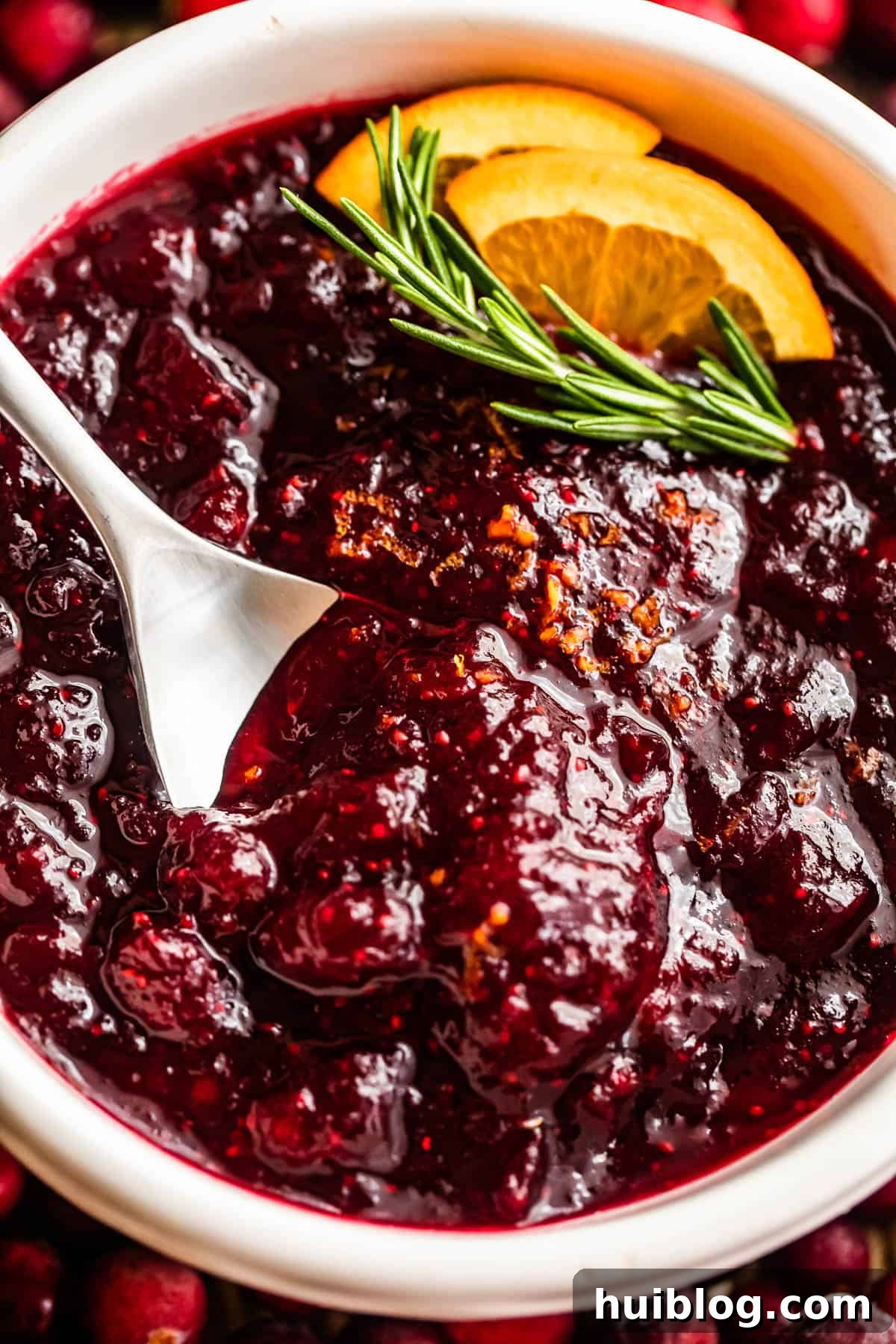 Overhead close-up shot of a spoon inside a bowl filled with homemade cranberry sauce, garnished with a rosemary sprig and fresh orange slices, ready for serving.