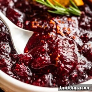 Side shot of a spoon inside a bowl filled with cranberry sauce, garnished with rosemary sprig and orange slices.