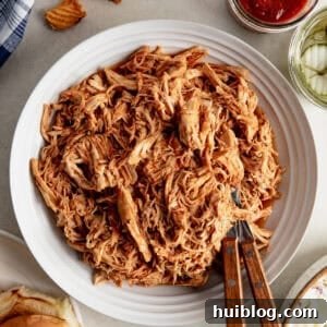 Shredded BBQ Chicken in a bowl with hamburger buns, pickles and sauce next to it.